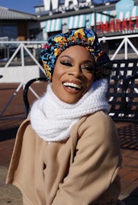a woman wearing a scarf and beanie smiles on a bench